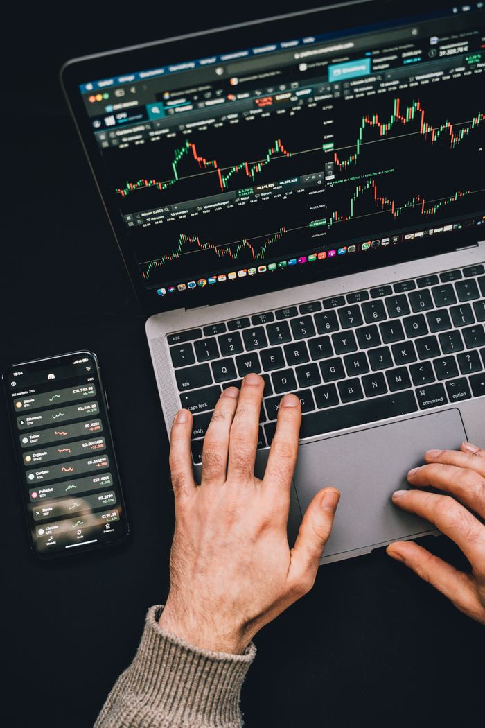about-us-01 Hands typing on a laptop with cryptocurrency charts and smartphone display in view, focusing on digital finance.